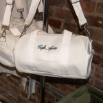 Load image into Gallery viewer, Beige duffel bag with 'Cafe Ayres' embroidery on a stand against a brick wall.