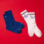 Load image into Gallery viewer, Two pairs of socks, one blue and one white with branding, on a red background.