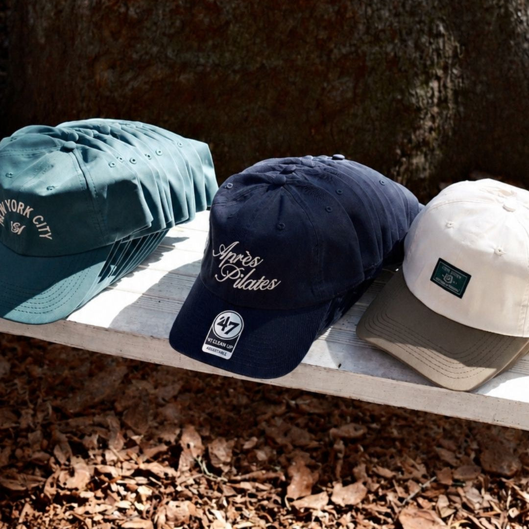 Three baseball caps displayed outdoors on a white bench in front of a large tree trunk, with dry leaves on the ground. The caps are teal, navy blue, and off-white, each with embroidered script text on the front and a visible '47 brand patch.