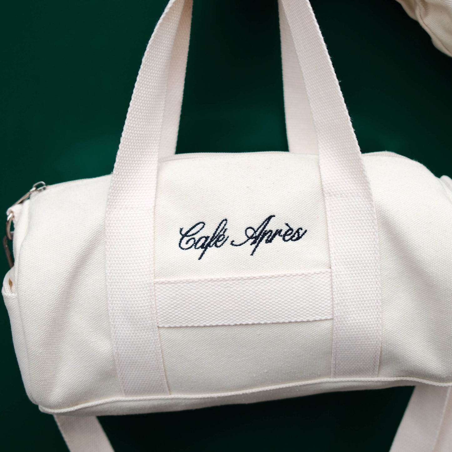 White duffel bag with 'Cafe Apres' embroidery on a dark green background