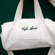 White duffel bag with 'Cafe Apres' embroidery on a dark green background