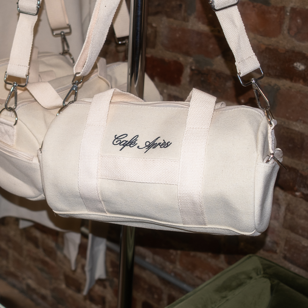 Beige duffel bag with 'Cafe Ayres' embroidery on a stand against a brick wall.