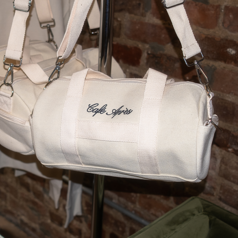 Beige duffel bag with 'Cafe Ayres' embroidery on a stand against a brick wall.