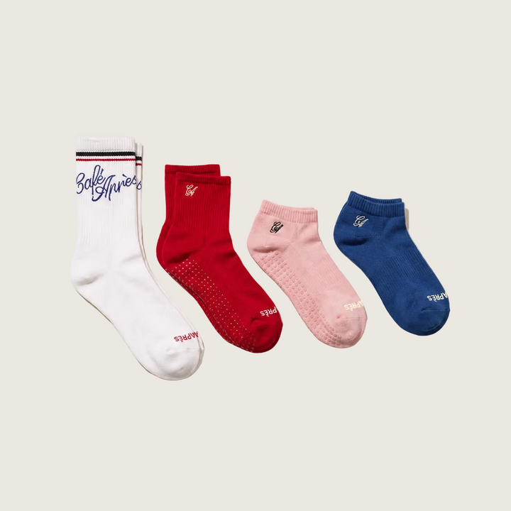 Four pairs of socks arranged diagonally on a light beige background: one white pair with navy script and thin black/red stripes, one red pair with a small white logo, one light pink pair with a small white logo, and one blue pair with a small white logo.
