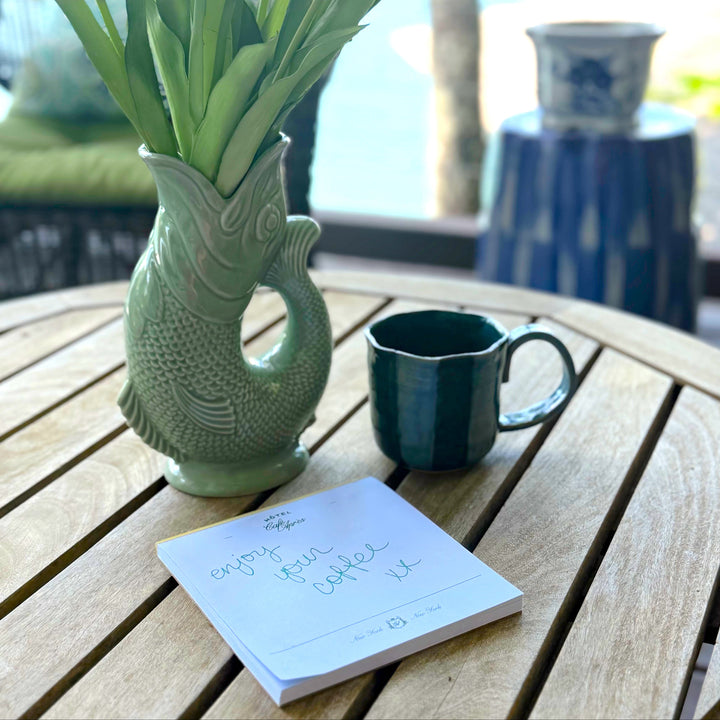 A light green ceramic fish-shaped vase filled with pink tulips sits on a round wooden table next to a dark green ceramic mug and a small handwritten notepad. The background shows a softly blurred outdoor patio setting with a blue decorative pot and green cushions.