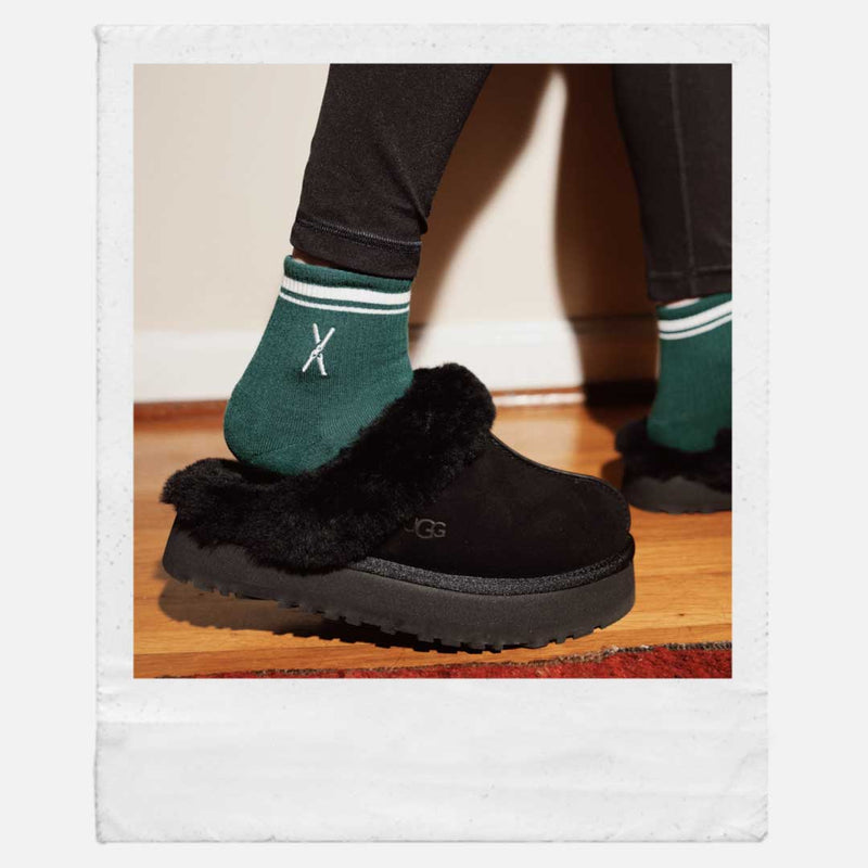Black slippers worn with green socks on a wooden floor