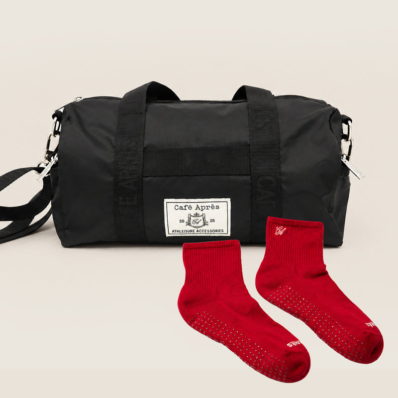 A black nylon duffel bag with a white 'Café Apres' brand patch on the front, displayed alongside a pair of red grip socks on a light beige background.