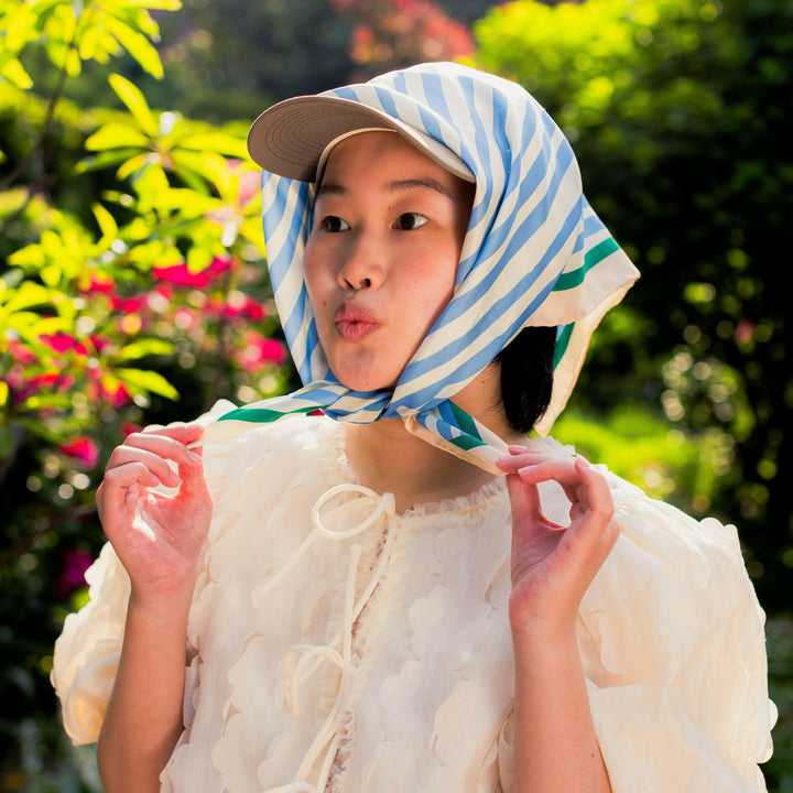 Woman wearing a striped headscarf and cap outdoors with greenery and flowers in the background